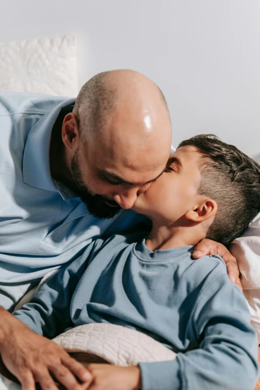 a man sitting on top of a bed next to a little boy, kissing each other, bald, headshot, enes dirig