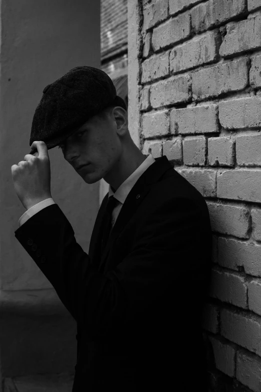 a man in a suit leaning against a brick wall, a black and white photo, inspired by Dmitry Levitzky, peaky blinders (2018), black hat, young male, ansel ]