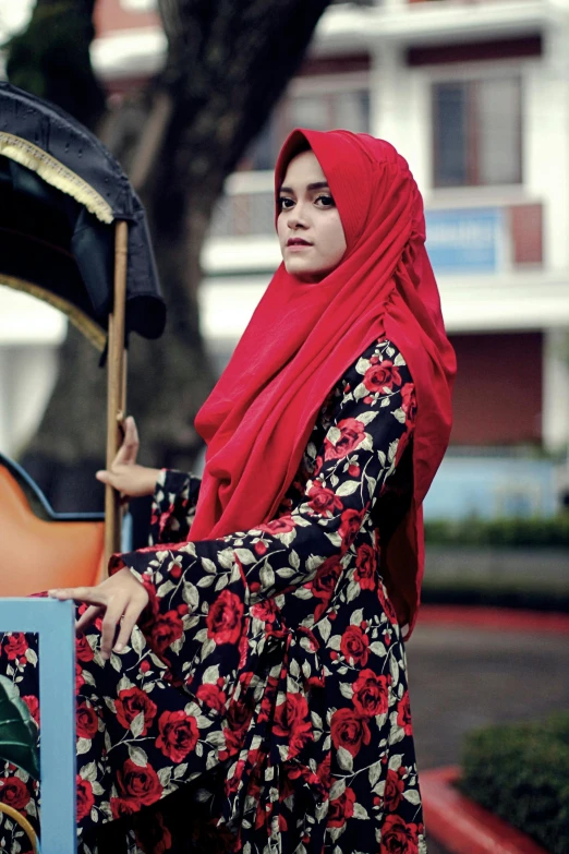 a woman in a red hijab stands next to a tuk - tuk, by Basuki Abdullah, pexels contest winner, hurufiyya, black and red silk clothing, floral, square, (beautiful) girl