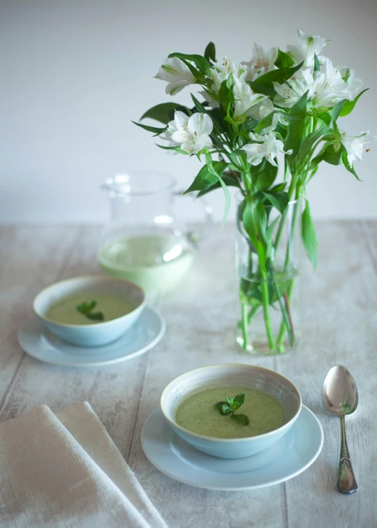 a table topped with two bowls of soup next to a vase of flowers, light green, food styling, floating. greenish blue, frog themed