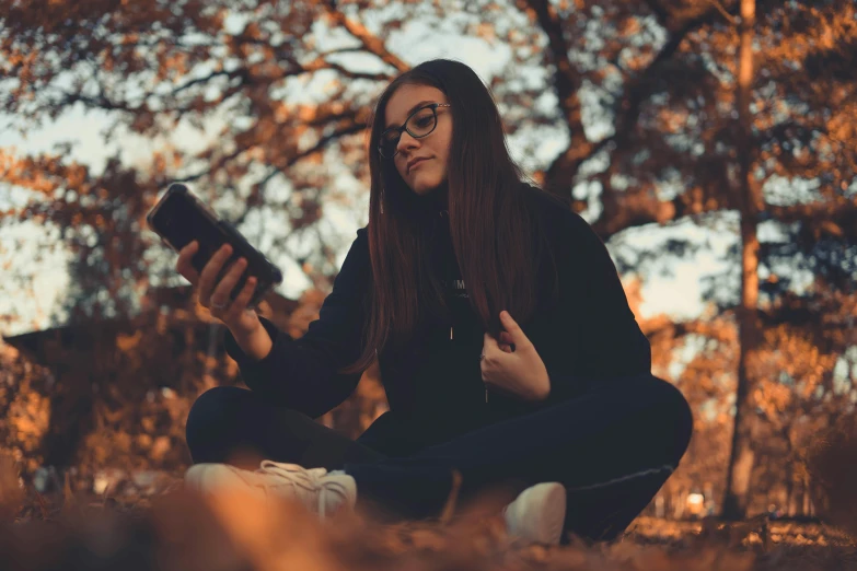 a woman sitting on the ground holding a cell phone, a picture, by Emma Andijewska, pexels contest winner, holding spell book, wearing black frame glasses, autumnal, profile pic