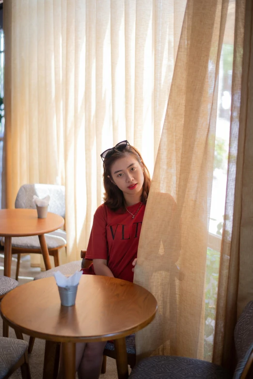 a woman sitting at a table in front of a window, a picture, unsplash, realism, red t-shirt, in tokio, sitting on a mocha-colored table, 15081959 21121991 01012000 4k