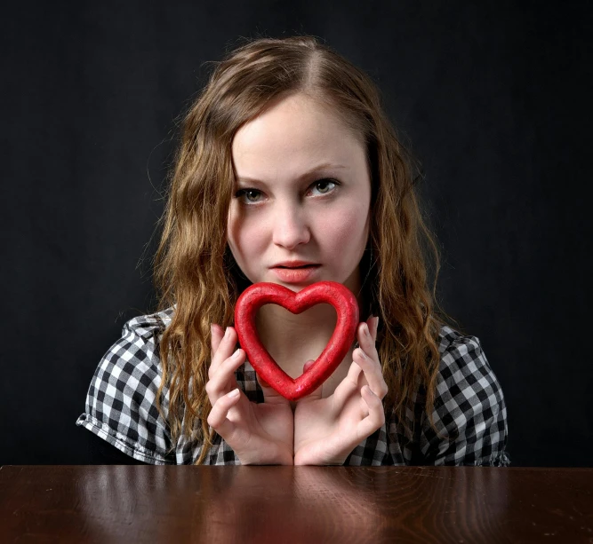 a girl holding a red heart in front of her face, a picture, by Adam Marczyński, pixabay contest winner, sitting on a table, square, studio shoot, beautiful symmetric