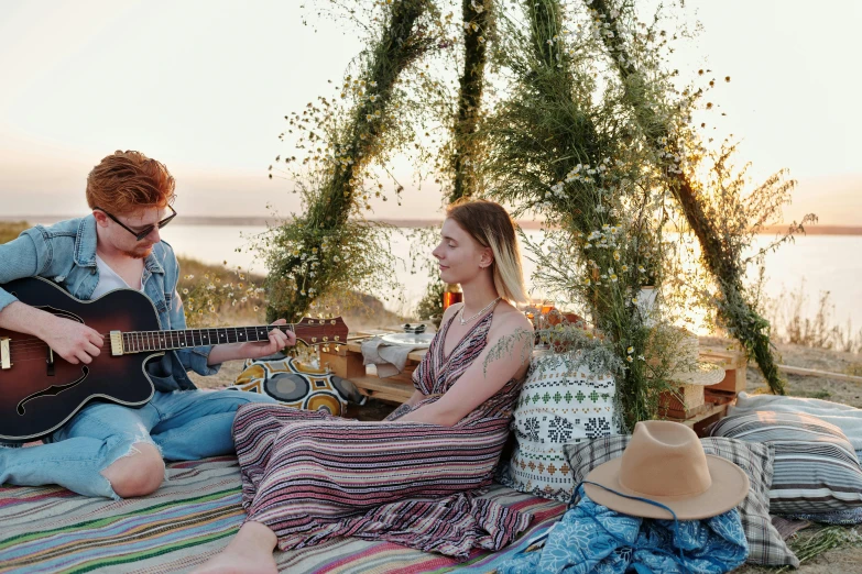 a man and a woman sitting on a blanket playing guitar, sydney sweeney, on the coast, flowers around, performing