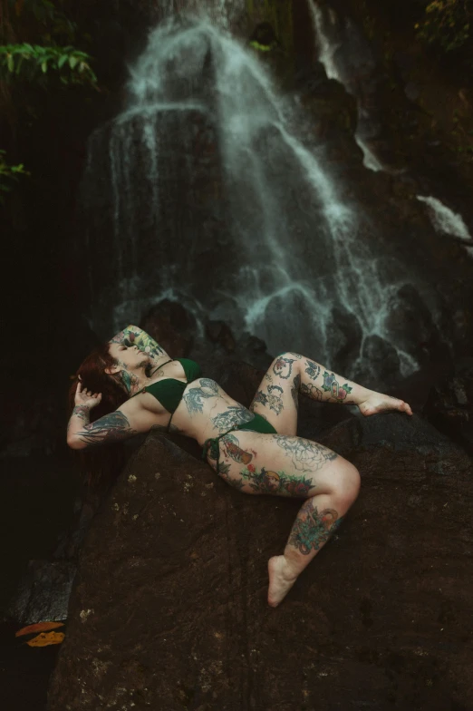 a woman laying on a rock in front of a waterfall, a tattoo, green skin, thicc, full body tattoo, full body shots