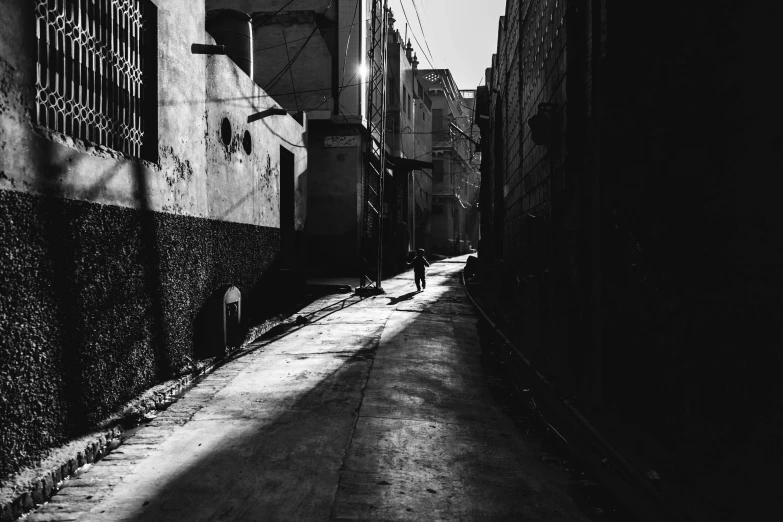 a black and white photo of a person walking down a street, by Ahmed Yacoubi, unsplash contest winner, old jeddah city alley, winter sun, late morning, kano)