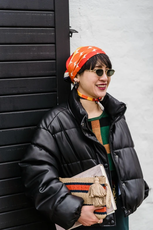 a woman leaning against a wall with a book in her hand, trending on pexels, visual art, wearing a bandana and chain, black and orange coat, beanie, asian features