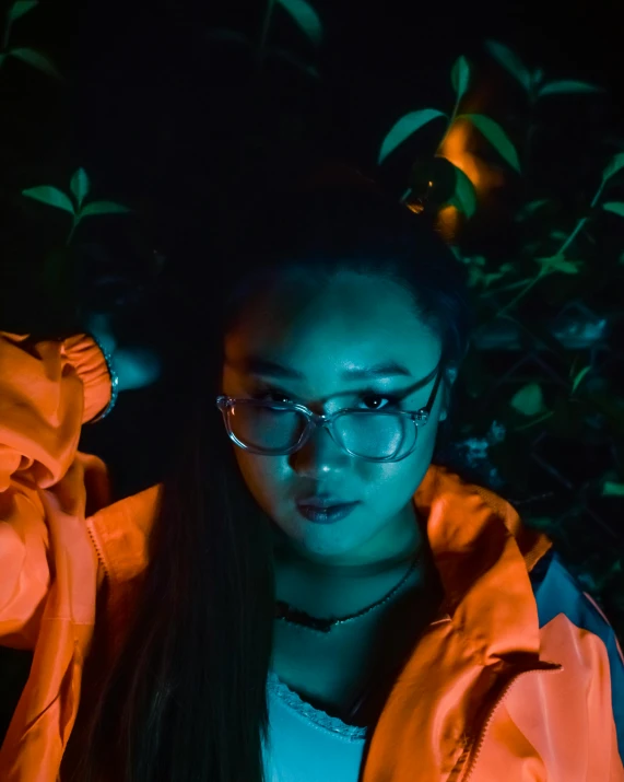 a woman wearing glasses and an orange jacket, inspired by Liam Wong, unsplash, visual art, bisexual lighting, looking down on the camera, ☁🌪🌙👩🏾, spooky photo