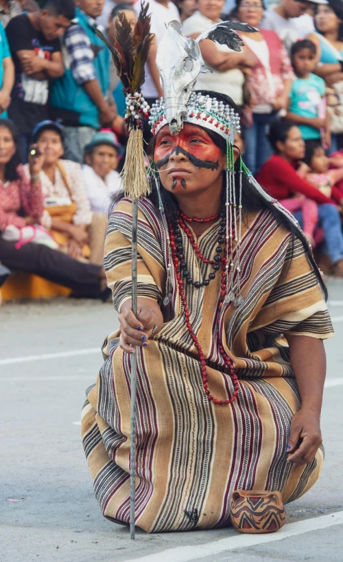 a man sitting on the ground in front of a crowd, by Delmer J. Yoakum, pexels contest winner, dau-al-set, she is dressed in shaman clothes, zigor samaniego, markings on her face, parade