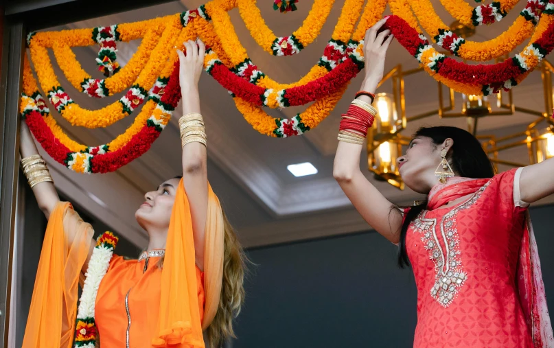 a couple of women standing next to each other, hurufiyya, flower decorations, hanging from the ceiling, orange and white, bollywood