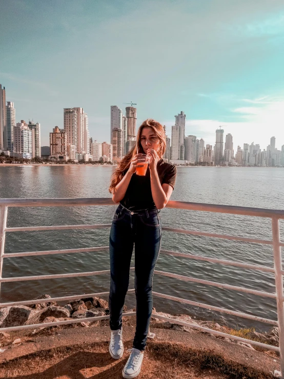 a woman standing in front of a body of water, a picture, with a city in the background, colombia, woman drinking coffee, avatar image