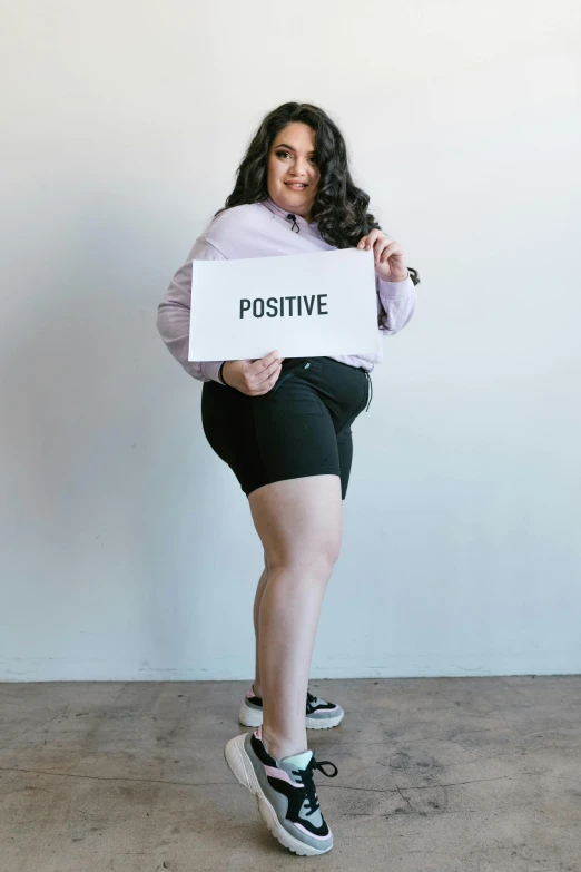 a woman holding a sign that says positive, by Olivia Peguero, curvy model, wearing black shorts, promo image, full body sarcastic pose