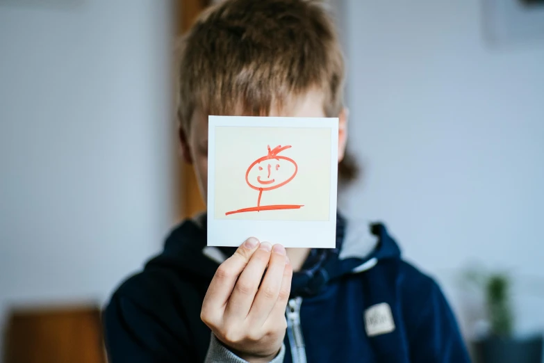 a boy holding up a piece of paper with a drawing on it, by Sebastian Spreng, pexels contest winner, square face, rated t for teen, headspace, polaroid picture