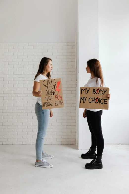 two women standing next to each other holding signs, trending on pexels, bodies, facing each other, profile image, girl standing