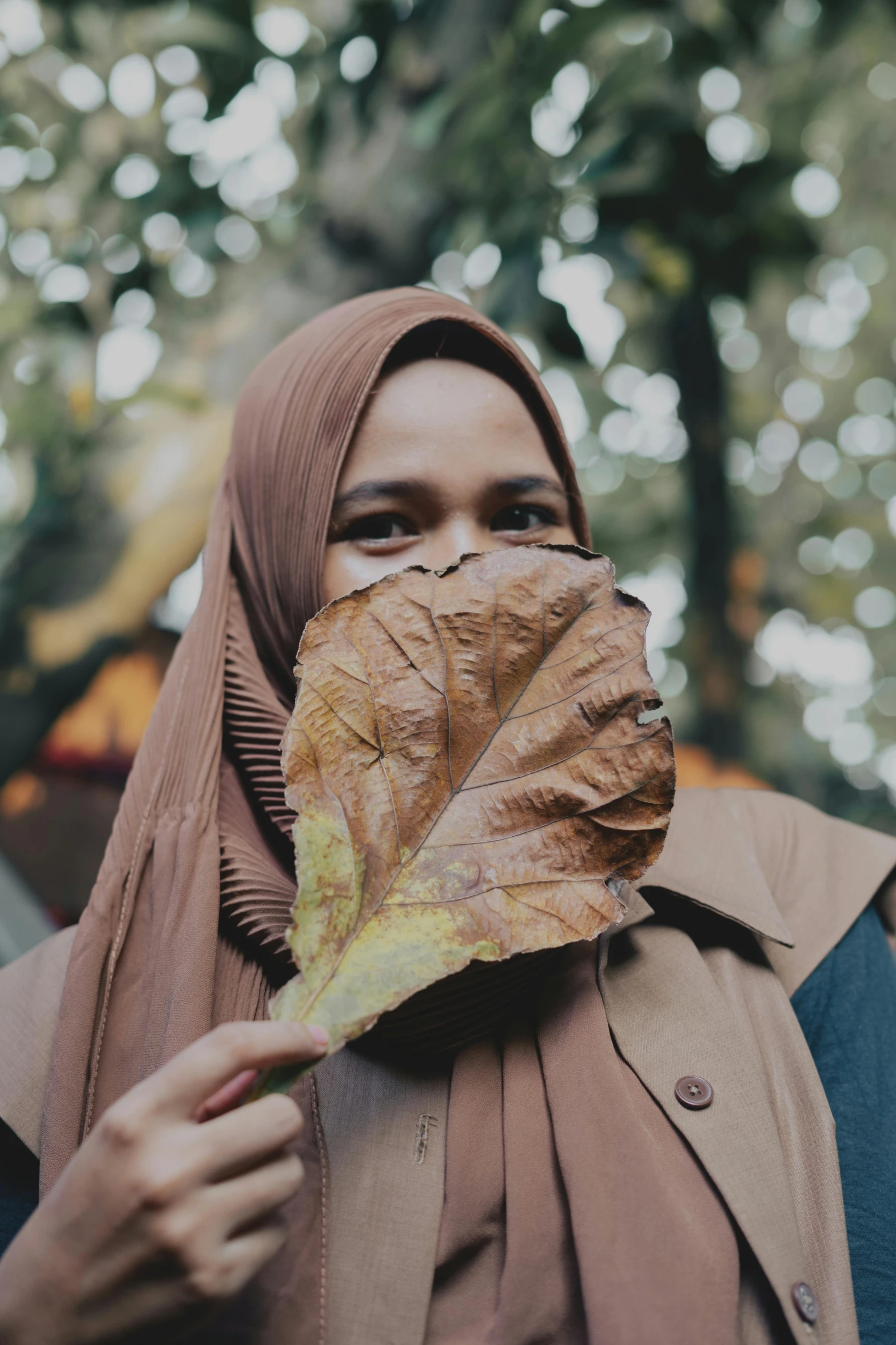 a woman holding a leaf in front of her face, an album cover, by Basuki Abdullah, pexels contest winner, hurufiyya, wearing a brown, bruh moment, ((portrait)), muslim