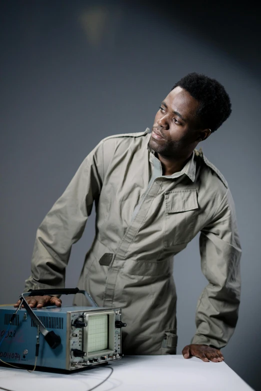 a man standing next to a radio on a table, an album cover, inspired by Edwin Georgi, yasuke 5 0 0 px models, kevin hart, wearing human air force jumpsuit, looking to the right