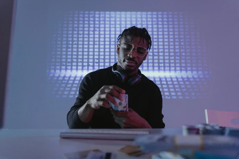a man sitting at a table in front of a projector screen, digital art, inspired by Ismail Gulgee, video art, purple liquid in cup glowing, like a cyberpunk workshop, offering the viewer a pill, shot with sony alpha 1 camera