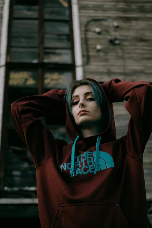 a woman in a red hoodie standing in front of a building, inspired by Elsa Bleda, trending on pexels, square face, maroon and blue accents, menacing pose, sport clothing