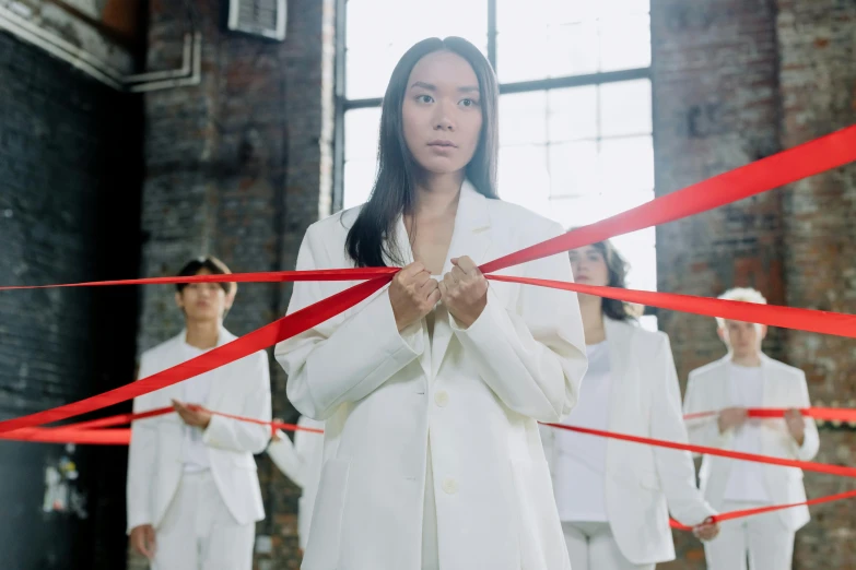 a woman holding a red ribbon in front of a group of people, inspired by Vanessa Beecroft, pexels contest winner, gutai group, wearing futuristic white suit, louise zhang, ( ( theatrical ) ), tailored clothing