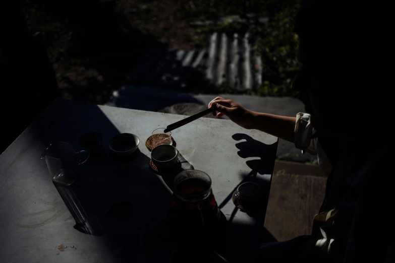 a person sitting at a table with a knife in their hand, unsplash, process art, assam tea garden setting, in the shadows, pouring, cliffside