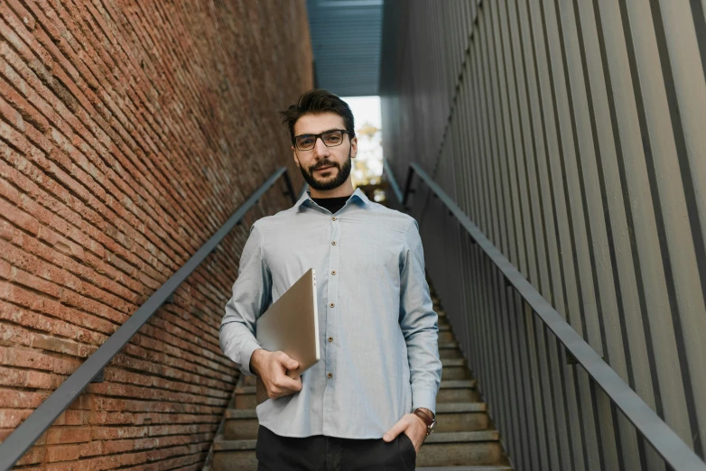 a man standing on a set of stairs holding a folder, pexels contest winner, computer nerd, jewish young man with glasses, emad mostaque, 555400831