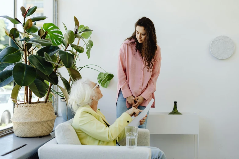 a woman standing next to a woman sitting in a chair, by Emma Andijewska, pexels contest winner, hurufiyya, health supporter, sitting on couch, on a white table, old and young