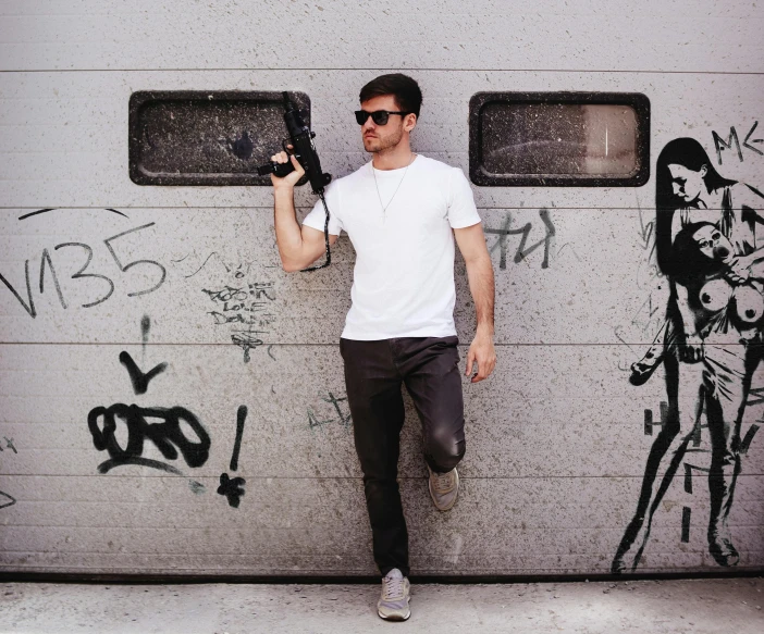 a man leaning against a garage door holding a camera, an album cover, by Bernie D’Andrea, pexels contest winner, graffiti, holding a blaster, man in white t - shirt, mid-shot of a hunky, maxim shirkov