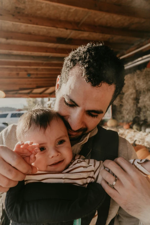 a man holding a baby in a sling, pexels contest winner, on a farm, clear julian lage face, hugging, square