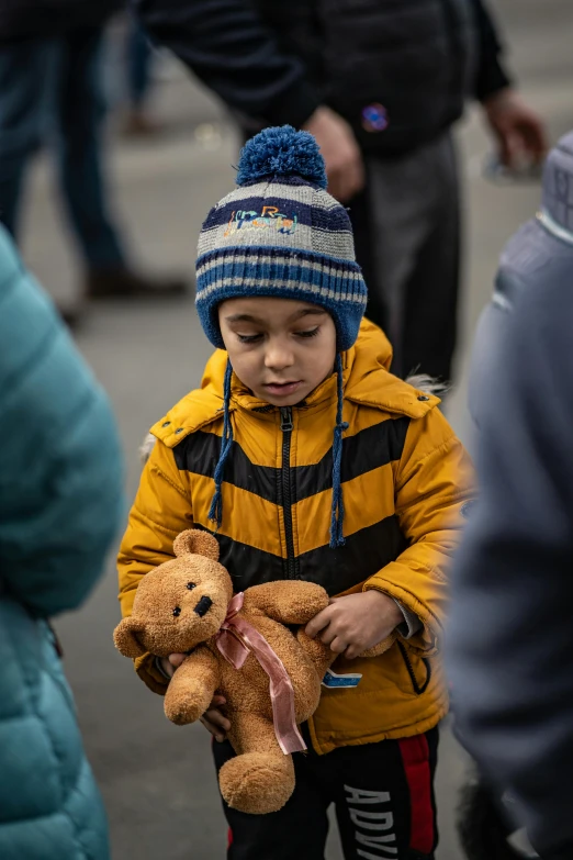 a little boy that is holding a teddy bear, by Adam Marczyński, pexels contest winner, refugees, square, beanie, wearing blue jacket