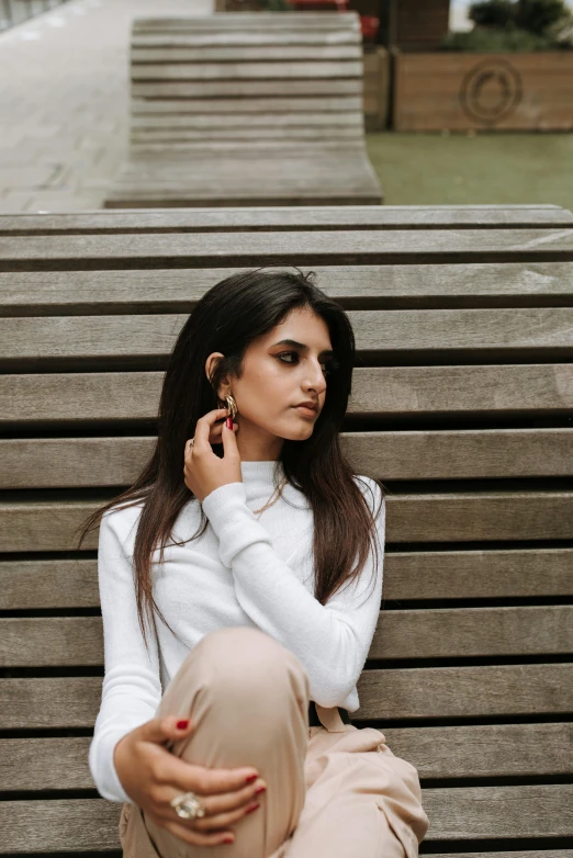 a woman sitting on a bench talking on a cell phone, trending on pexels, hurufiyya, in white turtleneck shirt, indian girl with brown skin, stoic pose, wearing an elegant outfit