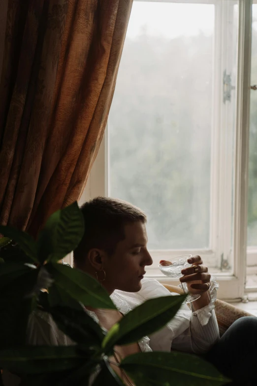 a man sitting on top of a couch next to a window, a picture, unsplash, delicate androgynous prince, is ((drinking a cup of tea)), ignant, slight haze