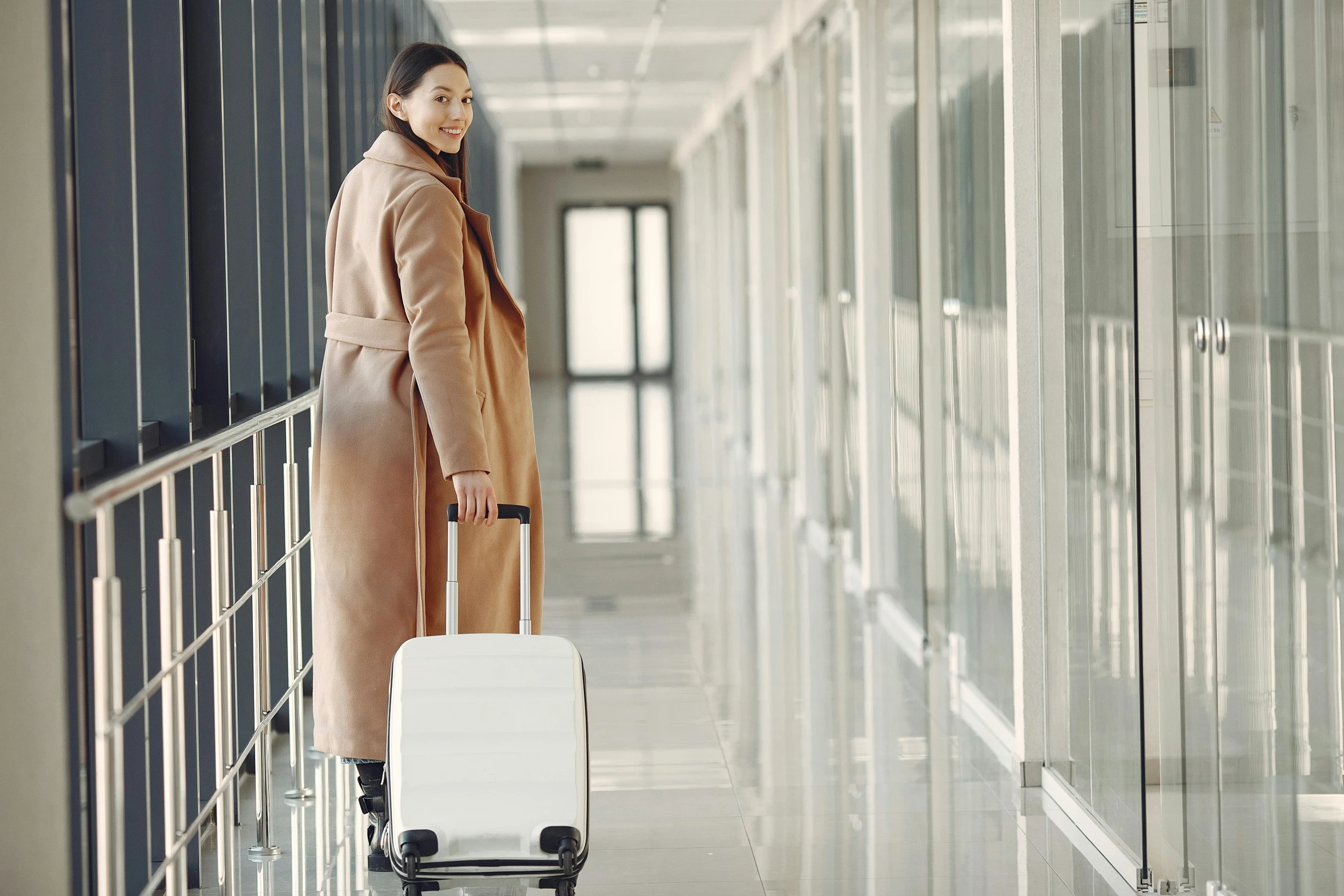 a woman standing in a hallway with a suitcase, happening, wearing a white winter coat, advanced technology, thumbnail, light brown coat