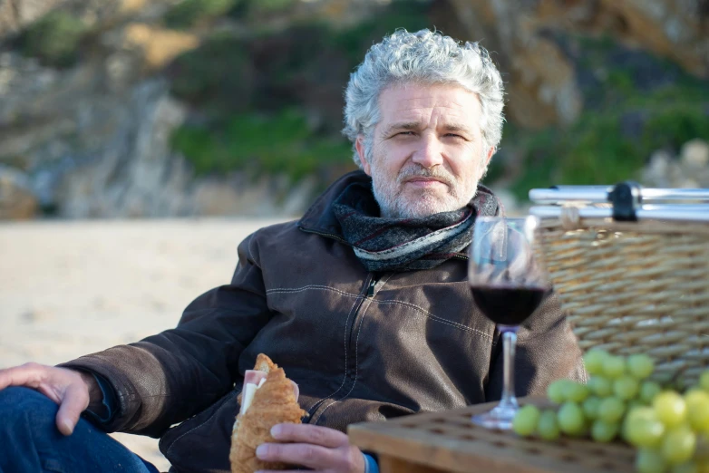 a man sitting at a table with a glass of wine, on the sand, grey beard, having a picnic, profile image