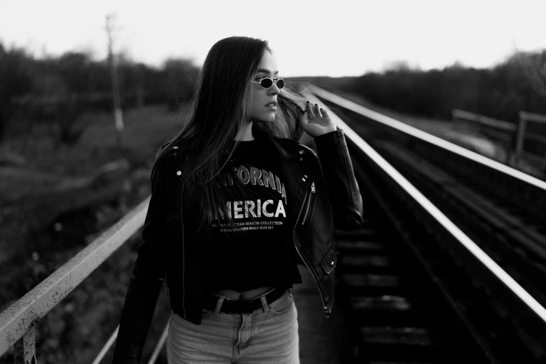 a woman standing on a train track talking on a cell phone, a black and white photo, by Emma Andijewska, pexels, graffiti, very aesthetic leather jacket, americana, girl with glasses, black shirt with red star
