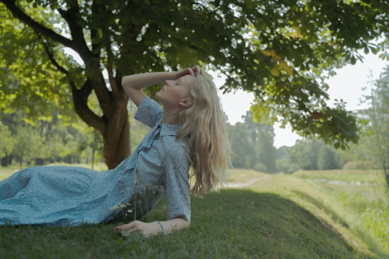 a woman in a blue dress laying on the grass, by Alice Mason, pexels contest winner, under the soft shadow of a tree, 4 k cinematic still, with long blond hair, the wind moves her shirt