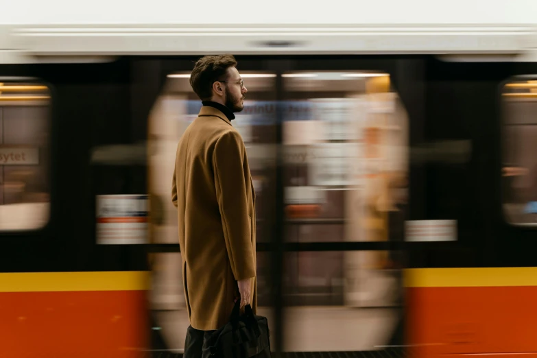 a man that is standing in front of a train, by Tobias Stimmer, pexels contest winner, black and orange coat, london underground tube station, light brown coat, thumbnail