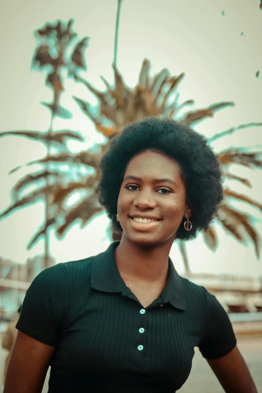 a woman standing in front of a palm tree, by Chinwe Chukwuogo-Roy, pexels contest winner, renaissance, dark short curly hair smiling, portrait androgynous girl, wearing a black shirt, ( brown skin )