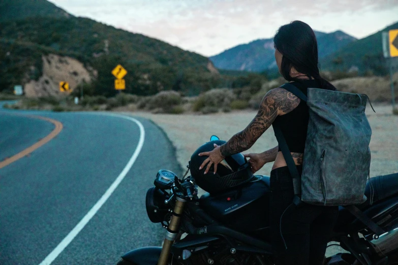 a woman sitting on a motorcycle on the side of the road, by Tom Bonson, pexels contest winner, symbolism, with a backpack, tattooed back, southern california, triumph