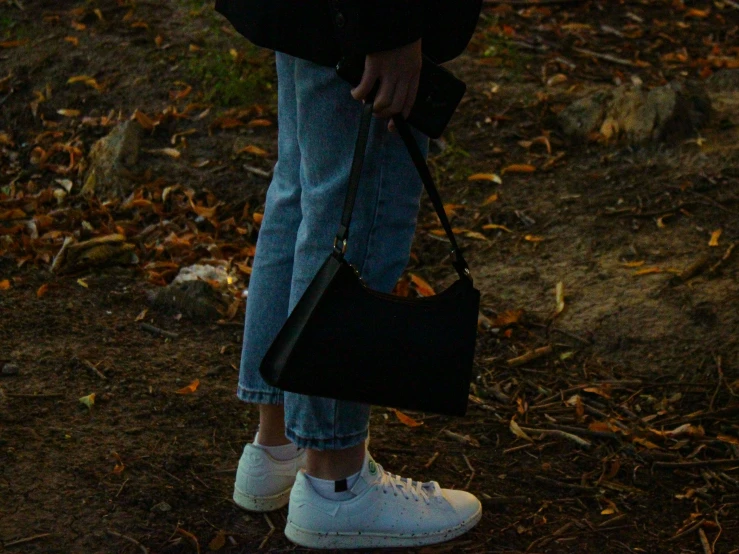 a person standing in a field holding an umbrella, wearing white sneakers, holding a leather purse, out in the dark, denim