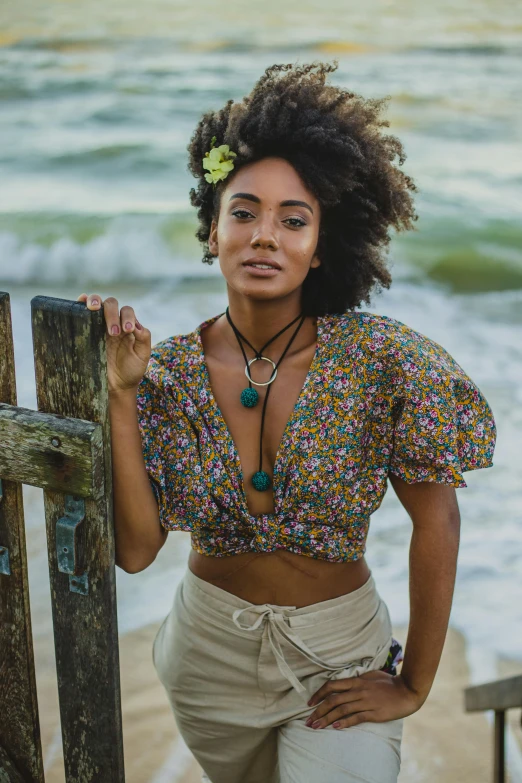 a woman standing on a beach next to the ocean, a portrait, trending on pexels, renaissance, wearing a crop top, wooden jewerly, afro made of flowers, promotional image