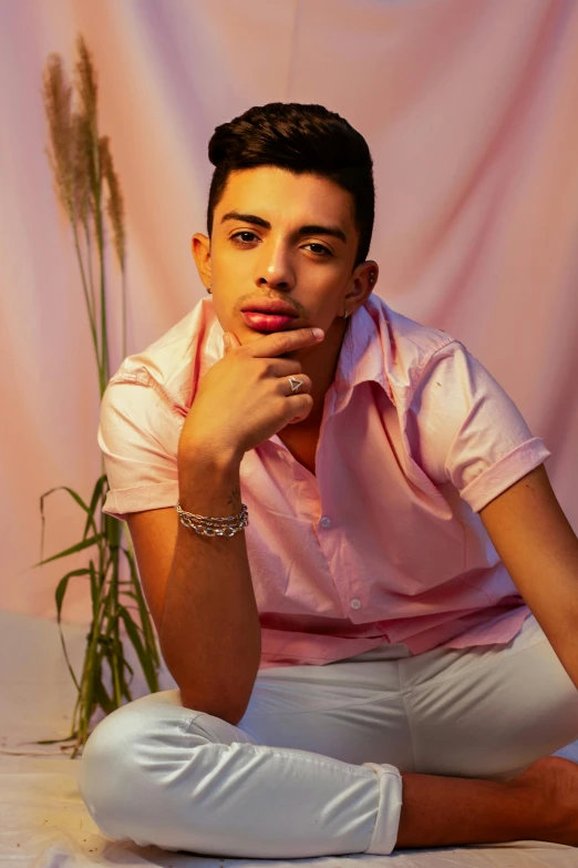 a man sitting on top of a bed next to a plant, an album cover, inspired by Carlos Berlanga, trending on pexels, pink shirt, androgynous face, middle eastern, crossed arms