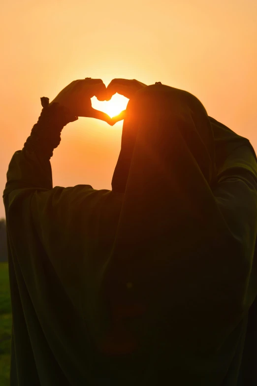 a person making a heart shape with their hands, inspired by Sheikh Hamdullah, pexels contest winner, hurufiyya, sun behind her, burka, profile image, of a lovely