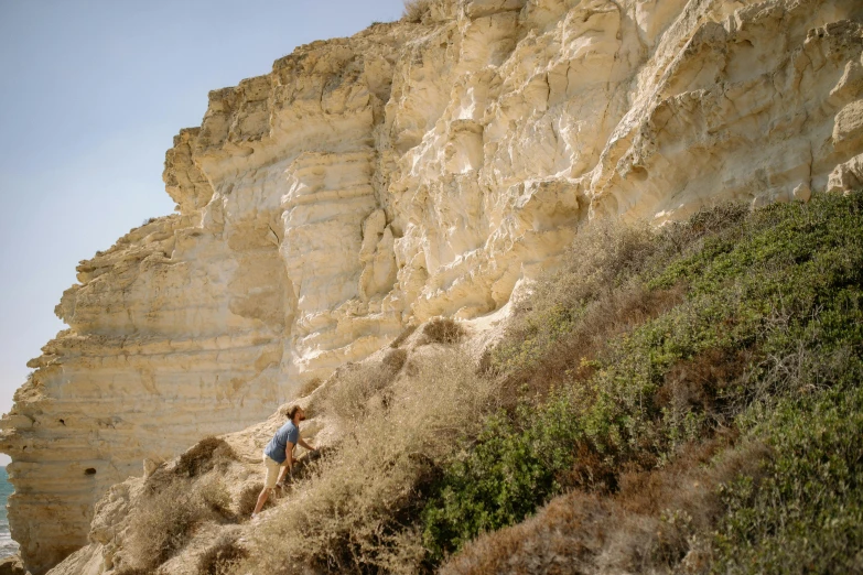 a man climbing up the side of a cliff, by Simon Marmion, unsplash, cyprus, girl walking between dunes, chalk cliffs above, ochre