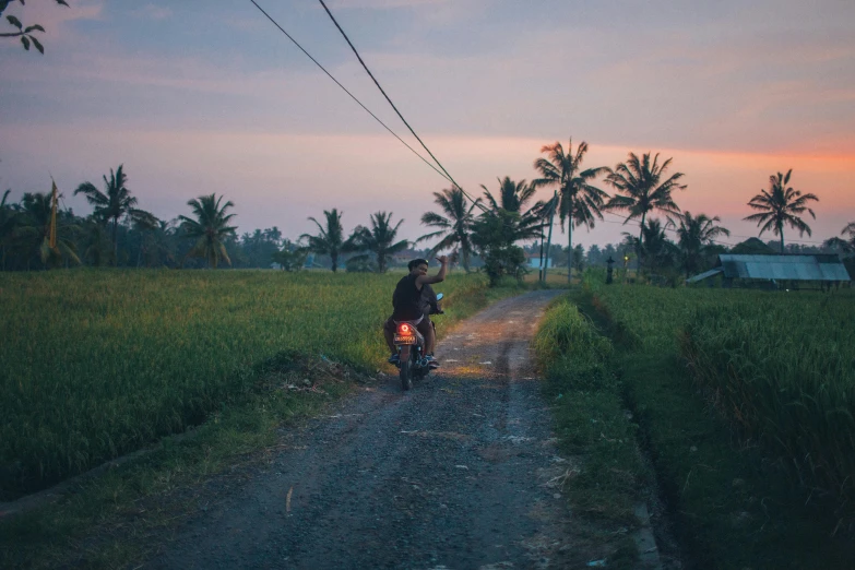 a man riding a motorcycle down a dirt road, inspired by Steve McCurry, pexels contest winner, sumatraism, spring evening, neighborhood, rice paddies, lights on