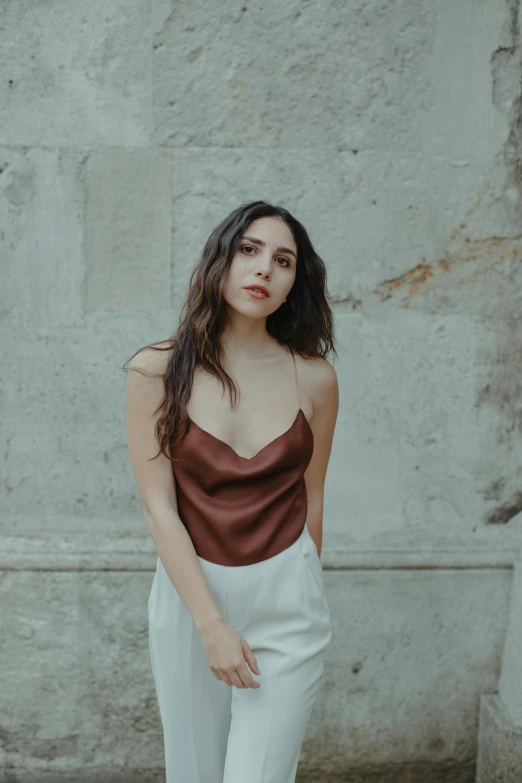 a woman standing in front of a stone wall, an album cover, inspired by Elsa Bleda, trending on pexels, renaissance, wearing a camisole, oona chaplin, brown clothes, white silky outfit