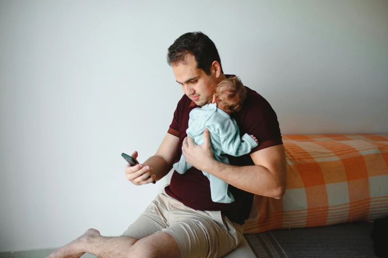 a man sitting on a bed holding a baby and looking at a cell phone, by Adam Marczyński, happening, struggling, brown, handheld, casually dressed