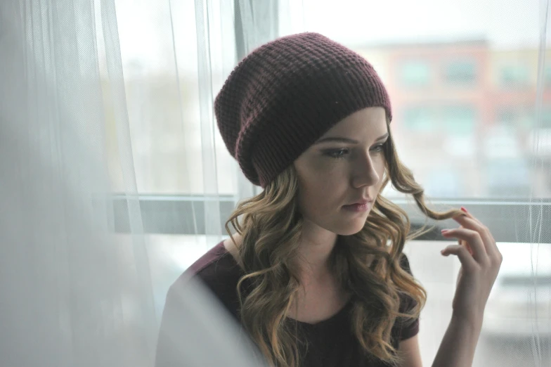 a woman in a hat looking out a window, an album cover, inspired by Elsa Bleda, photorealism, wearing a beanie, brown reddish hair, devon cady-lee, maroon