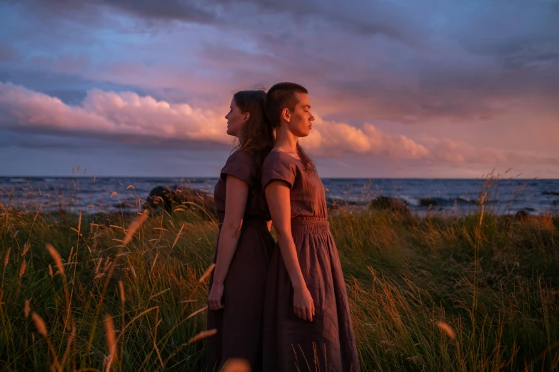 a couple of women standing on top of a grass covered field, by Else Alfelt, unsplash, romanticism, beside the sea, [ theatrical ], evening lighting, eleven/millie bobbie brown