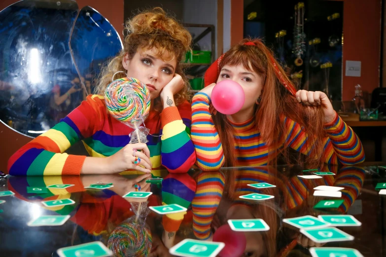 a couple of women sitting next to each other on a table, an album cover, kitsch movement, lollipops, youtube thumbnail, playing board games, colourful clothing