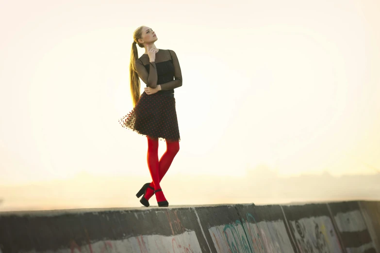 a woman standing on top of a cement wall, an album cover, inspired by Elsa Bleda, pexels, renaissance, in red velvet stockings, with a ponytail, sunrise light, concert